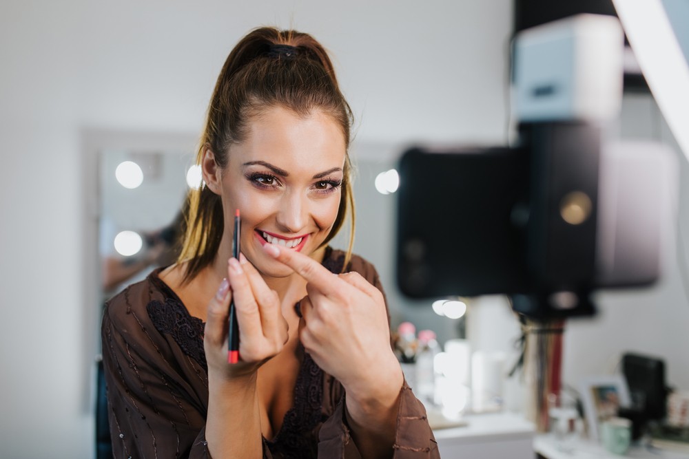 Blonde woman applying lip liner in front of a phone that's recording her. A vanity can be seen in the background.